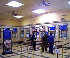A room with a high yellow ceiling with fluorescent lights and ceiling fans. It has a crisscross black pattern on a tan floor, with ropes and stanchions creating space for a line. Along the walls are teller windows with bronze grilles. The logo of the United States Postal Service is on many displays in the image
