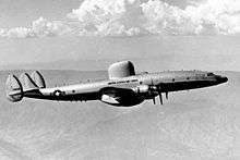 A monochrome photograph of a large aircraft flying above dimly-visible land, below white clouds, the aircraft showing multiple engines, three vertical tail surfaces and a large, rounded radar structure on its upper surface above and between the wings