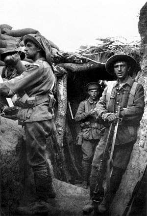A black and white photograph of men wearing military units in a trench. One man stands on a parapet looking away to the left, while others behind him stare into the camera