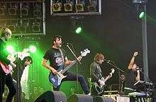 Five musicians playing, from left to right, guitar, vocals, guitar/vocals, bass, keyboard/vocals. They play with green lights in the background and large unused spotlights above their heads.