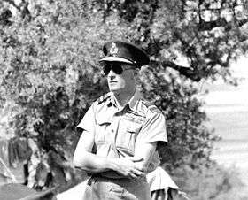 Outdoor half portrait of man in light-coloured military uniform with peaked cap, wearing sunglasses