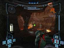 View of volcanic caverns; an enemy with a jetpack shoots a green ray at the player, whose weapon (a large cannon) is visible in the corner of the screen. The image is a simulation of the heads-up display of a combat suit's helmet, with a crosshair drawn onto the enemy's location and two-dimensional icons relaying game information around the edge of the frame.