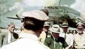 We see the back of MacArthur's head. Truman in a dark suit and light hat stands at a microphone. Behind him stands four mean in shirts and ties. In the background are some onlookers and a sleek metal but propeller driven airliner. Another aircraft lies parked on the runway.