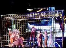 A giant cage, in front of which are gathered a number of people. Among them, a blond woman in brown jacket, pants and boots, stands on top of one of the rods and watches two men dancing.