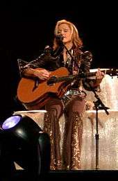 A blond woman sitting on a block of hay. She is playing a guitar and singing in front of a standing microphone. She has short hair and wears grey colored cowboy clothes.