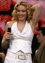 The front profile, from the waist up, of a middle-aged blond woman. She is wearing a white, sleeveless coat and white pants. Her hair is parted in the middle and is in locks around her face. She is holding a microphone in her right hand while her left hand is placed behind her head. She is smiling looking down. Behind her a video screen is red.