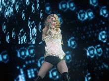 Image of a blonde woman. She is wearing a yellow sparkling corset with black shorts and black knee-high boots. She has her left arm pointing forward. She's standing in front of a blue backdrop,