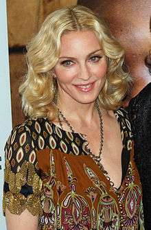 A blonde Caucasian female in a calico blouse and a silver chain smiles while looking to the left and slightly downward in front of a flesh-toned backdrop.