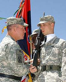 two men wearing army combat uniforms, one holding an M-4 as part of a color guard, the other placing a combat patch on the other's shoulder.