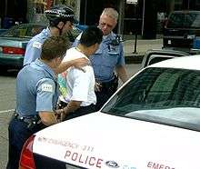 three police officers surround a man in a tee shirt who is handcuffed