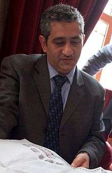 A man wearing a suit with short black and white hair, Mansoor Al-Jamri, reading a paper