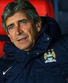 A photograph of a gentleman in his 60s. He is sitting on a red side bench. He is wearing a navy blue hooded jacket with a Manchester City crest on the left breast and a white logo of the Nike sponsor on the right breast.