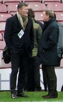 Two men wearing mostly dark coloured clothing stand at the side of a football pitch, hands in pockets, staring past end other.