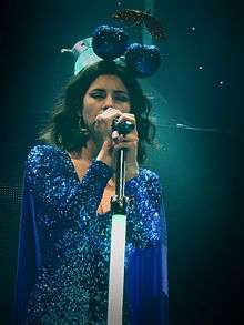 A light-skinned brown-haired woman is wearing a long-sleeved sparkling blue dress and a headpiece shaped like a pair of sparkling blue cherries, and is seen singing into a microphone.
