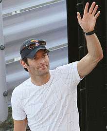 A man in his mid-thirties waving his left hand. He is wearing a white T-shirt and a dark blue hat with sunglasses on top
