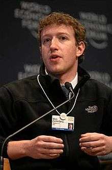 Waist high portrait of man in his twenties, looking into the camera and gesturing with both hands, wearing a black pullover shirt that says "The North Face" and wearing identification on a white band hanging from his neck