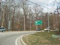 A road at a roundabout in a rural area with a sign to the left reading north Maryland Route 2 upper right arrow Maryland Route 408/Maryland Route 422 and a green sign to the right reading Maryland Route 2 north Annapolis straight
