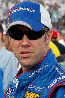 A head and shoulders portrait of a man in his late thirties. He is wearing black sunglasses and facing near to the camera. He is wearing a blue and white baseball cap which displays the CitiFinancial logo at the front. He is also wearing blue racing overalls, upon the collar which the CitiFinancial is embroidered and on the shoulders and body. Logos for Ford, DeWalt and Sparco are also displayed.