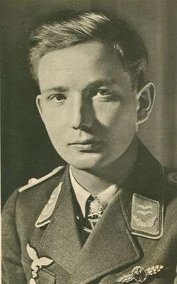 The head and shoulders of a young man, shown in semi-profile. He wears a military uniform with an Iron Cross displayed at the front of his shirt collar. His hair is dark and short and combed to his right, his nose is long and straight, and his facial expression is emotionless; looking into the camera.