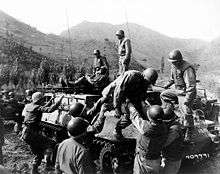 A group of soldiers are surrounding a tracked vehicle that carries two injured soldiers. One of the injured is getting off with the help of two soldiers while another is lying on top of the vehicle.