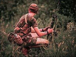 A young man with white skin and sandy-coloured hair squats in a field of tall grass, his body pointing to the viewer's right and his face looking away from the photographer. He is wearing a camouflage pattern cap, T-shirt and shorts and carrying numerous munitions and pieces of equipment. He holds between his hands a camouflage-painted FN FAL battle rifle, which is pointed upwards and away from him, its butt resting on the ground.