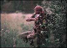 A young man with sandy blond hair, wearing brown and green Rhodesian-pattern military camouflage and a mesh scarf around his head, squats behind the cover of a large bush as he takes aim with a camouflaged FN FAL battle rifle. He is carrying a large backpack and has liberally applied dark camouflage cream to his skin.