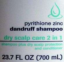 White slightly curved plastic with a green line drawing of a drop, the text "pyrithione zinc dandruff shampoo" in dark blue with "dry scalp care 2 in 1" in green, then a blue line. Underneath it is "shampoo plus dry scalp protection and conditioner" in smaller green type, and finally "23.7 FL OZ (700 mL)" in larger blue type.