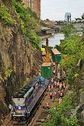 A red, blue and silver locomotive on tracks in a narrow gap between two tall rocky cliffs. To its right are a group of men wearing safety vests and hard hats. Behind it are some more of the large green metal containers seen in the picture above, a tall yellow crane and a small yellow truck. In the background is the train station area shown above, with the bridge that photograph was taken from visible at the upper right