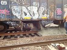 Large gravel, grayish in color, pours from a vent in the undercarriage of a blue hopper car with graffiti over dark brown gravel on the track below it