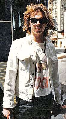 Upper body of a 26-year-old man. He is standing with his arms alongside. He has collar length brown hair. His light coloured jacket is opened to reveal a white T-shirt with an obscured image and printing. He wears dark glasses and the left side of his face is partly in shadow. Over his right shoulder is the entry way to a building, with other buildings nearby.