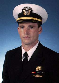 Color photograph of Michael Murphy, a U.S. Navy officer, wearing a military dress uniform. There is a blue background behind him and he is wearing a gold Navy Seal Trident, two blue and green striped ribbons, one red and yellow striped ribbon and gold parachute insignia wings below the ribbons.