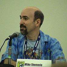 A bald man with a beard speaks into a microphone. A plaque with the Comic-Con logo on the table reads "Mike Stemmle".