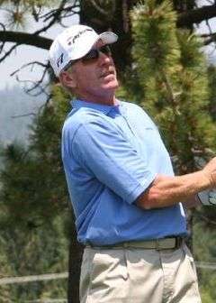 A white male with a receding hairline looks on after swinging at a golf ball with a club. He is wearing a sky blue polo shirt, golf pants, and a white cap.