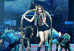 A teenage female with long, brunette hair grabs a microphone as she stands before a golden luggage cart. She is dressed in a black tank top, black hot pants, black leather boots and a blue jean vest. In the background, people ducking surround her and a DJ uses his equipment.
