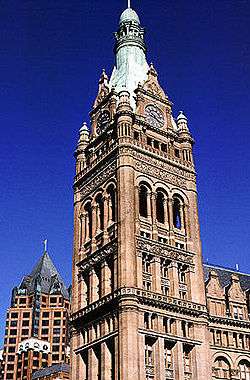 Ground-level view of a large, brick tower with several architectural niches and columns and a tapering, copper roof topped with a spire; a clock are visible on the tower near its roofline.