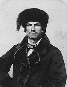 A black-and-white photograph of a man with Native-American features wearing a thick fur coat, a bow tie, and a fur hat.