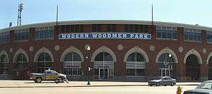 A large brick stadium with many round windows on the bottom and many rectangle windows in groups of four line the top of the stadium. The words Modern Woodmen Park are displayed above the door