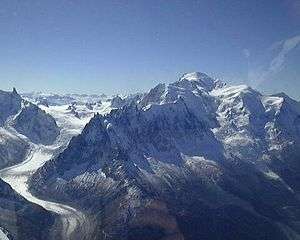 Photograph of a towering snow-covered mountain at right, a glacier at left, and a clear blue sky.