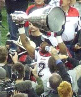 A player in a white uniform with red trim and a black number 2 on his chest raises a large silver trophy over his head.