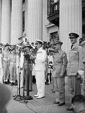 A man in a white uniform stands in front of a microphone on the steps of a building, surrounded by men in an array of uniforms.