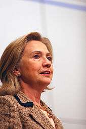 Informal pose of Clinton, in which she is wearing a beige wool jacket and facing toward the right. The angle at which Clinton is photographed is from her bottom right side.