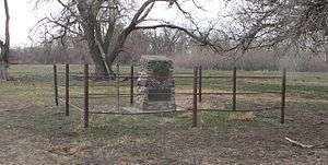 Stone monument about 3 feet high, on flat ground near wooded creek
