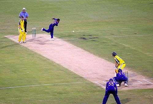 A bowler in a blue uniform bowls a white cricket ball to a batsman dressed in a yellow uniform and dark-blue helmet.  An umpire in a light blue shirt and cream, wide-rimmed hat watches, along with another batsman, at the other end of the pitch.  A wicketkeeper is kneeling close behind the batsman receiving the ball, and a slip fielder is standing behind and to his left, with his hands on his knees.