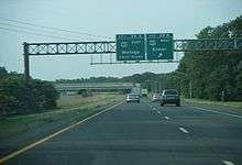 A four-lane freeway in a wooded area approaching an interchange. Two green signs stand over the highway with the left one reading exit 39A U.S. Route 40 east Malaga next right while the one on the right reads exit 39B U.S. Route 40 west Elmer with an arrow pointing to the upper right.