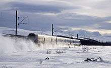 A four-car multiple unit running across a snow-covered plain with mountains in the background