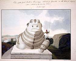 A huge bull, with large eyes, carved out of stone and ornamented with many necklaces, is seated on a stone altar built atop a hill and overlooking the plains behind. The bull's left front paw is extended out; its right, folded under. An adult man, shown in the picture, is dwarfed by comparison.