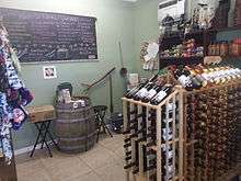 A room with green walls and tile floors with a rack of wine in the front, and a wine barrel and chalkboard in the back.