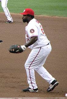 A man in a white baseball uniform with a red baseball cap and black glove plays in the field.