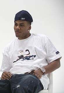 An African-American man sits and poses for a photo, wearing a white shirt, blue jeans, and baseball cap.