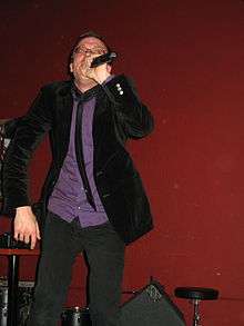 Photograph of a man in a purple shirt and dark suit, holding a microphone.
